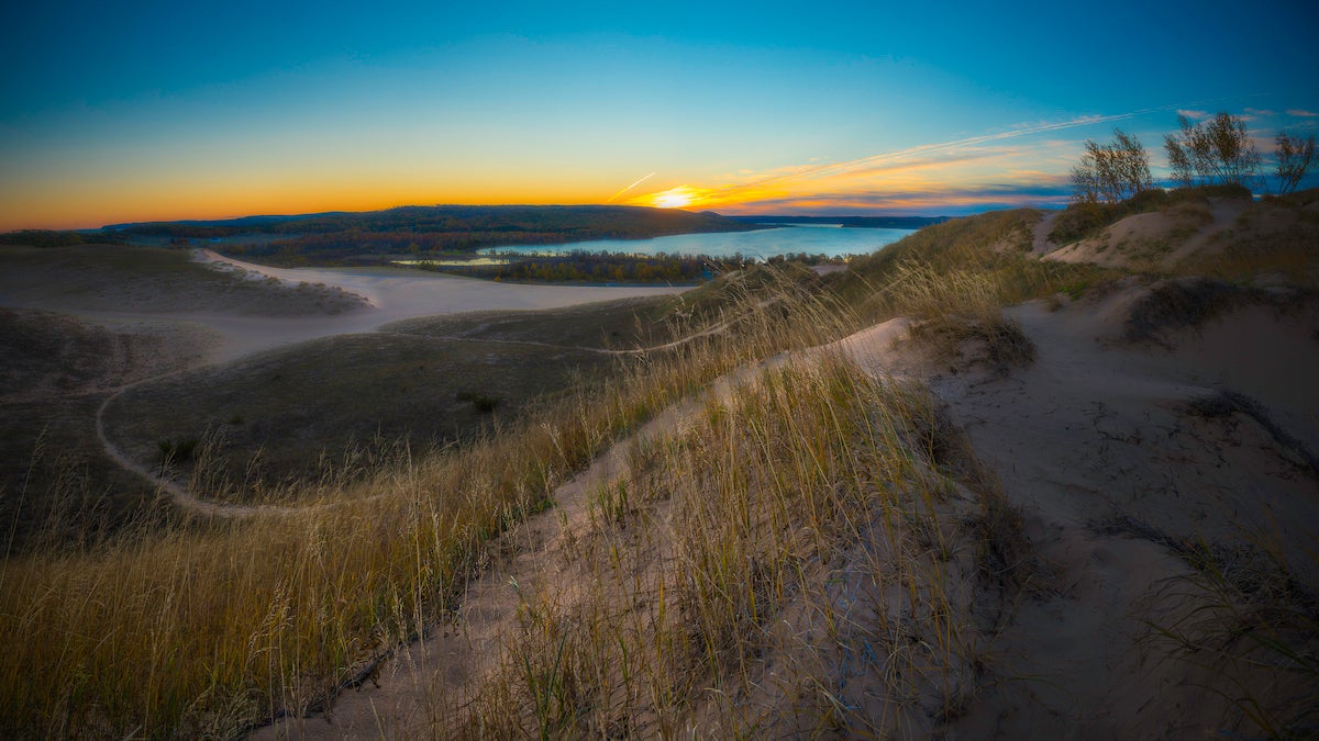 The sun rises over Glen Lake from the top of the Dune Climb in Sleeping Bear Dunes, as light and shadow play in the foreground.