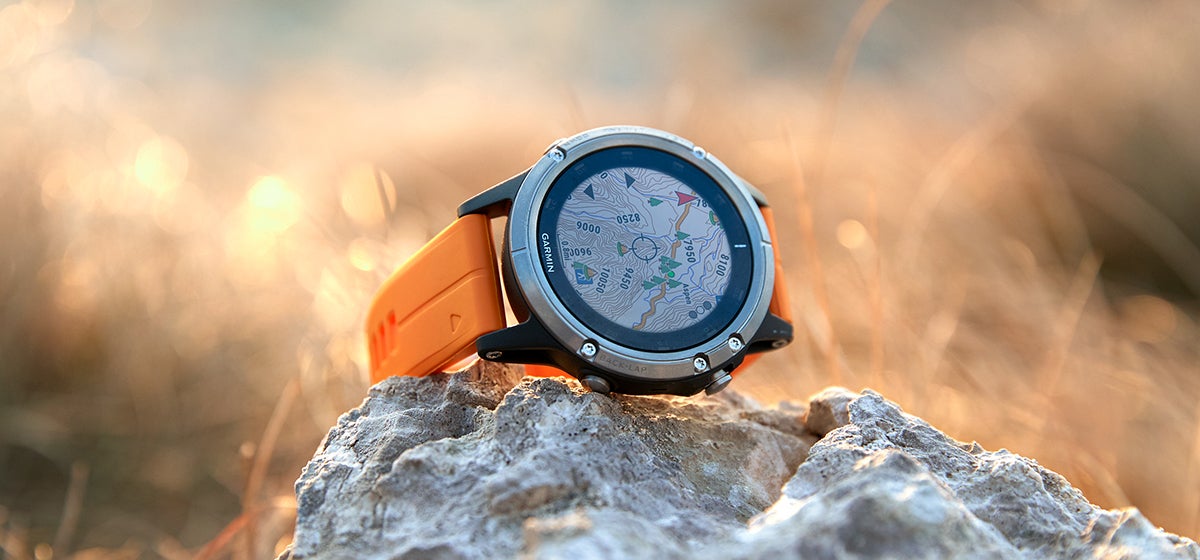 "Garmin fenix smartwatch with an orange band sits on a rock with sunshine and grass in the background."