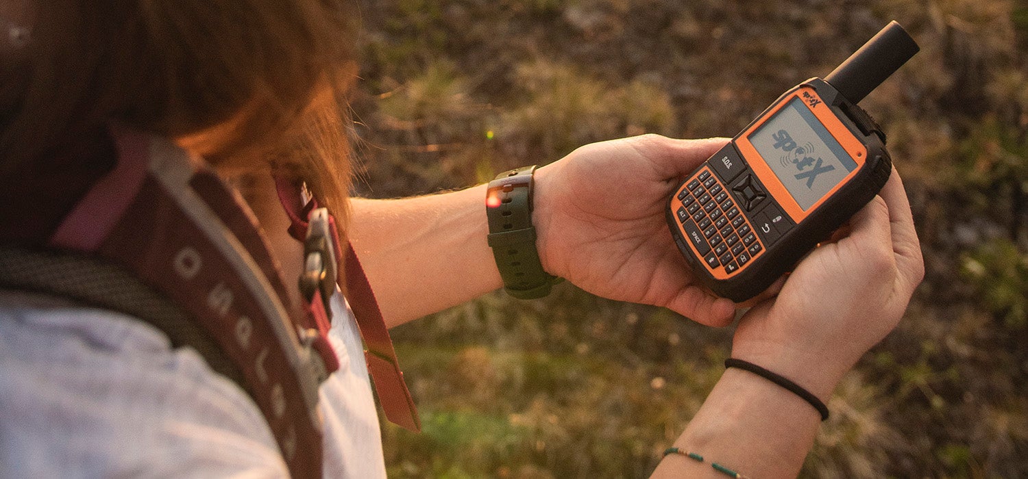 "White woman wearing Osprey backpack holds a black and orange SPOT X two-way satellite messenger device in her hands above the grass."