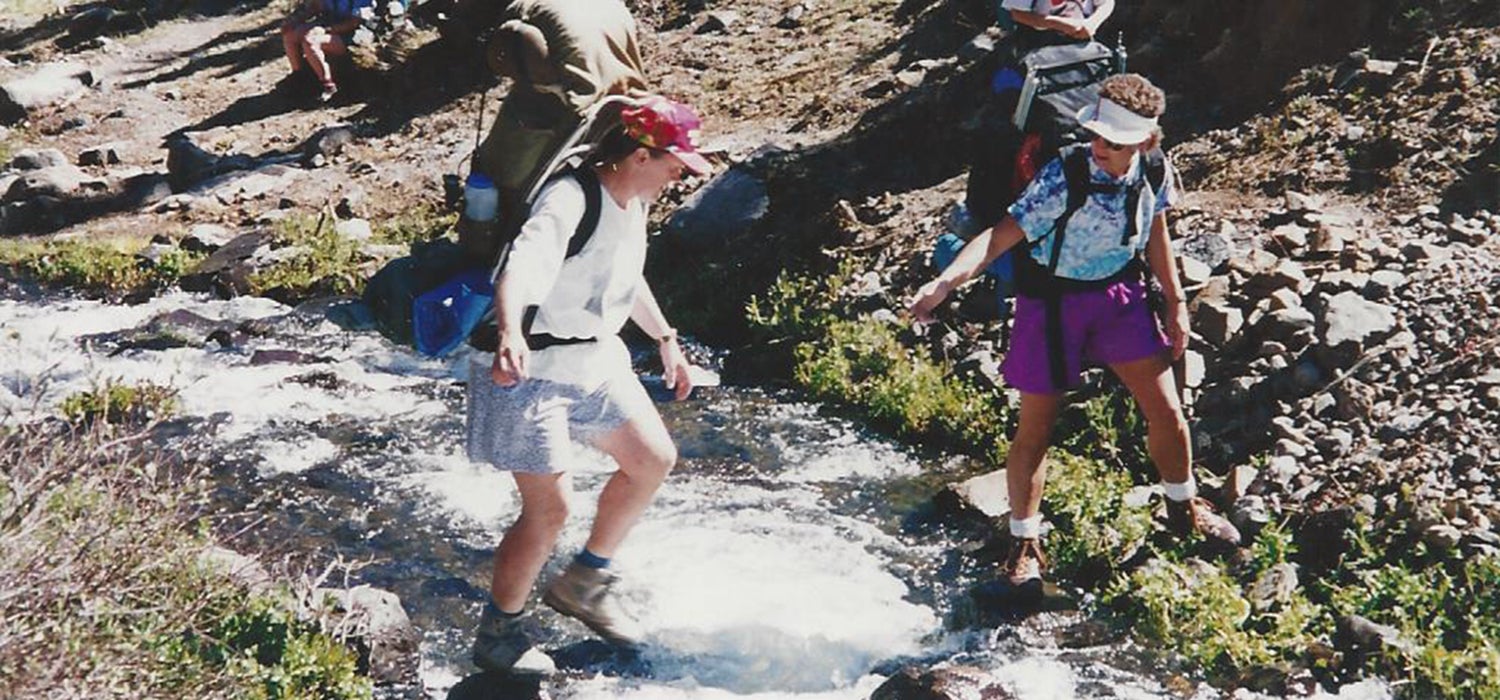 "Two women in 90s-style hiking gear (large flowy nylon shorts, baggy t-shirts, bright patterns) work together to cross Horn Stream."