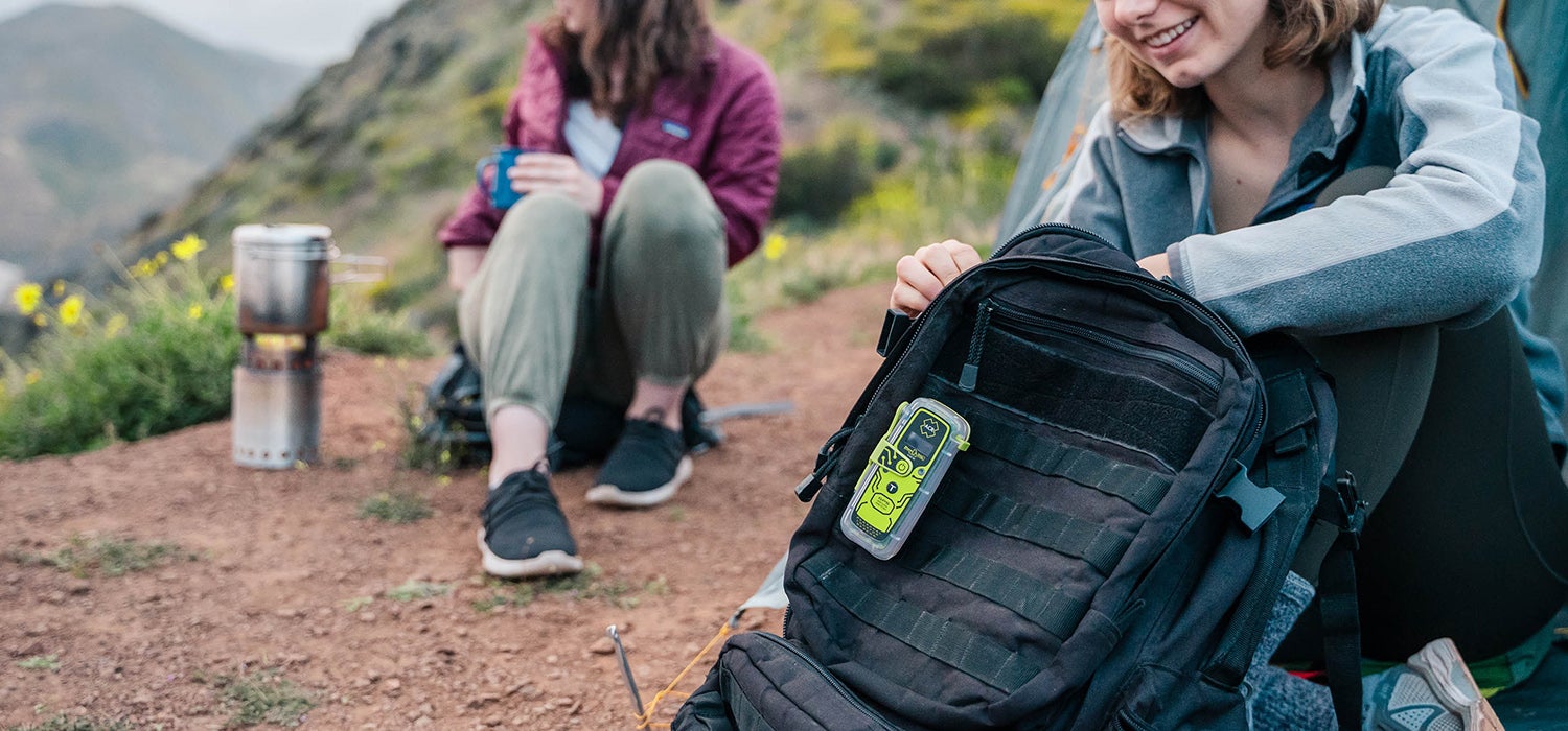 "Two women sit on a red dirt trail with grassy mountains in the background colored with yellow flowers. A black backpack is in focus with a yellow ACR&hellip;"