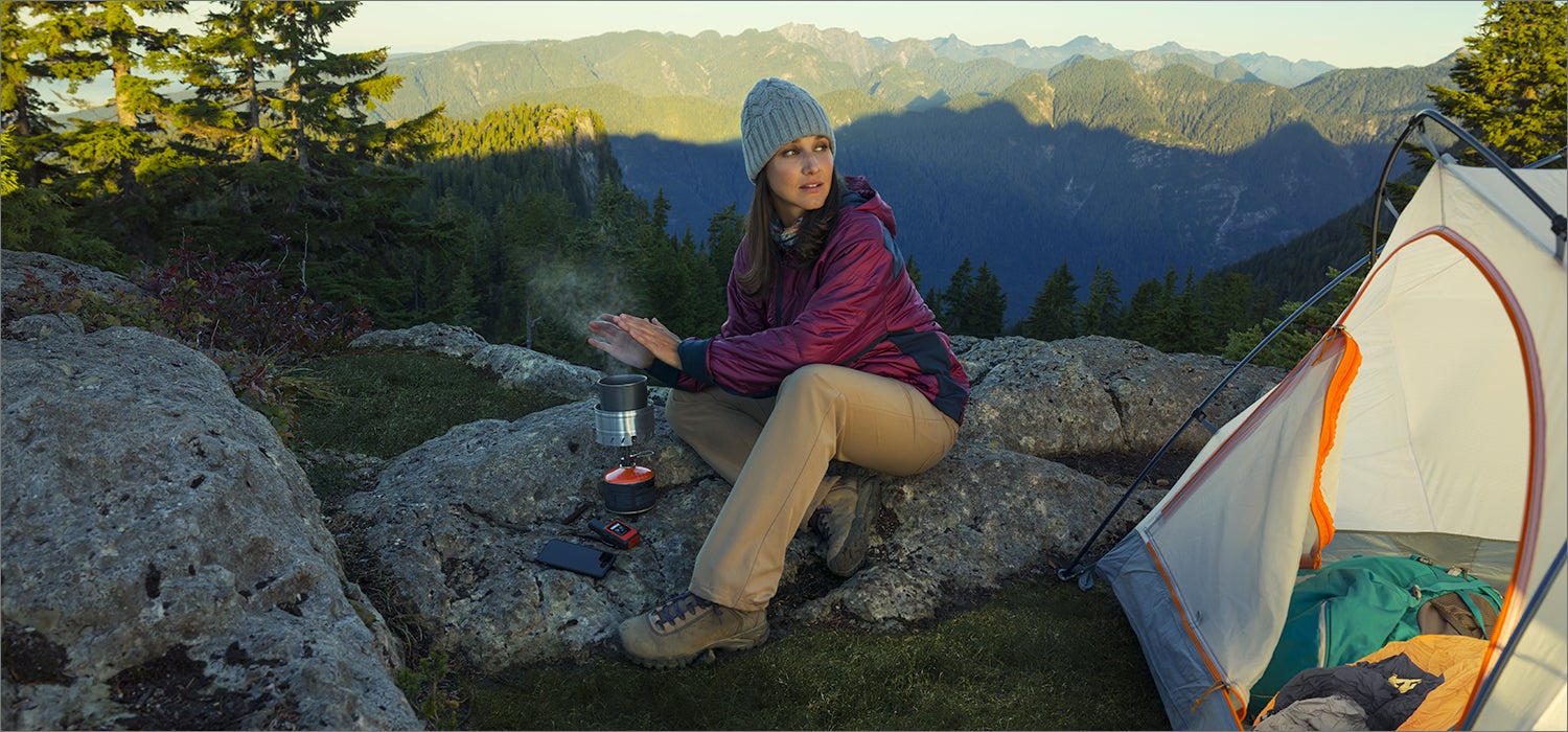 "Woman sits on a rock by a tent with mountain skyline in the background."