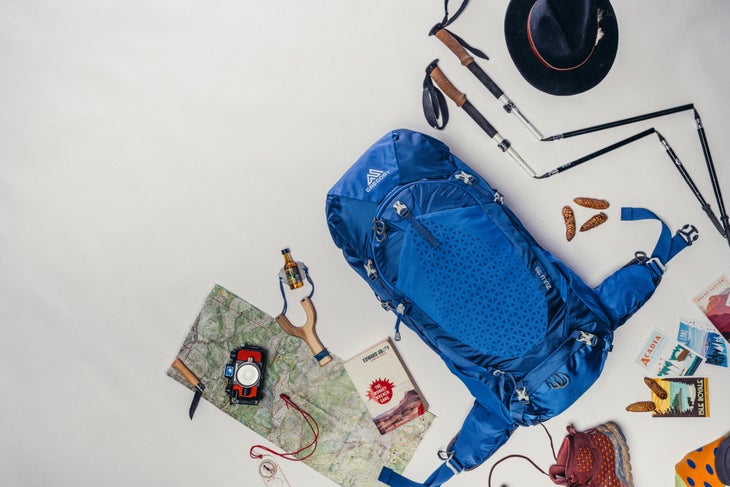 GMP_S19_Zulu_Enviro-168 Blue Gregory pack sits on a white background with supplies surrounding it such as a black felt hat, trekking poles, a slingshot, map, camera,…