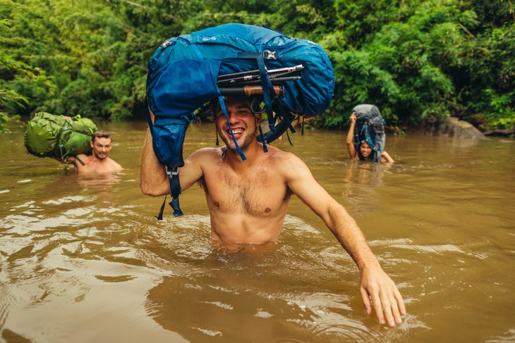 GMP_S19_ZuluJade_Hawaii-0453 Three people cross a muddy river in Hawaii laughing and holding their Gregory backpacking packs over their heads.