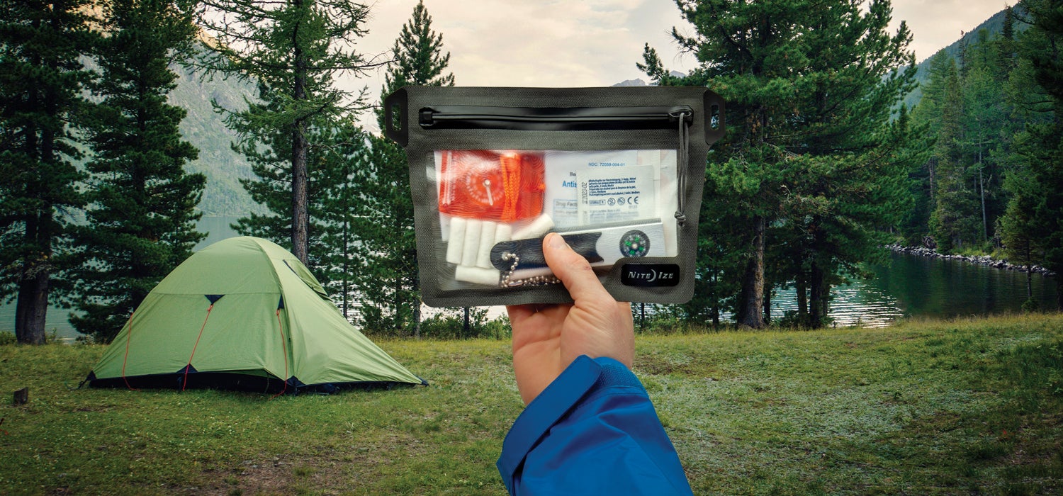 "White hand with a blue sleeve holds up a Nite Ize RunOff Dry Bag with a tent, lake, and evergreen trees in the background."