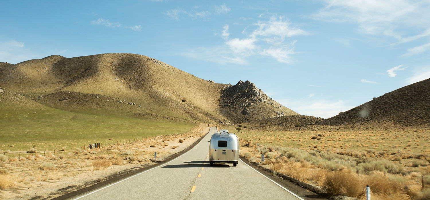 "Silver Airstream RV drives along straight road toward hills with green grass and brown shrubs alongside the the road."