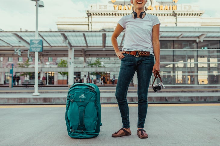 White woman stands on train platform holding camera with jade colored Gregory Proxy backpack on the ground beside her.