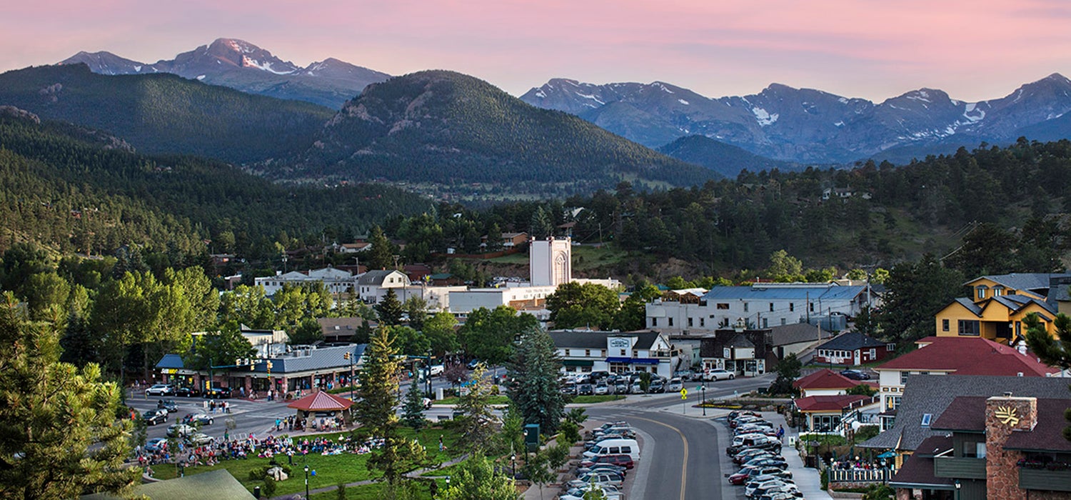 "Estes Park downtown is in the foreground with tree covered mountains making up the background below a pink sky."