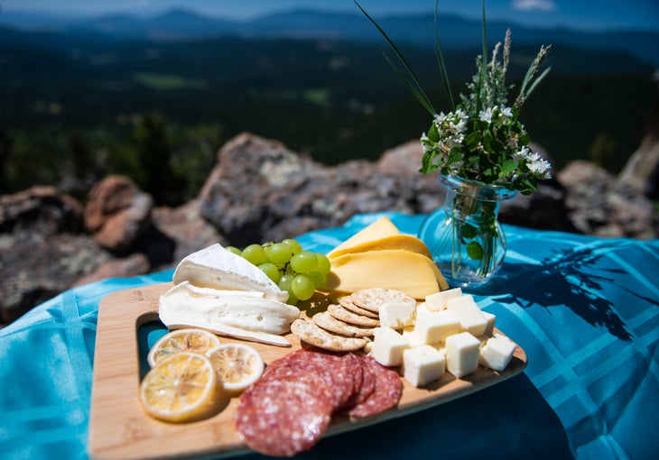 20190710_ViewRanger_100 Charcuterie board of salami, brie cheese, crackers, gouda, and cheddar cubes sit on a teal table clothe with a flower vase next to it. Rocks and…