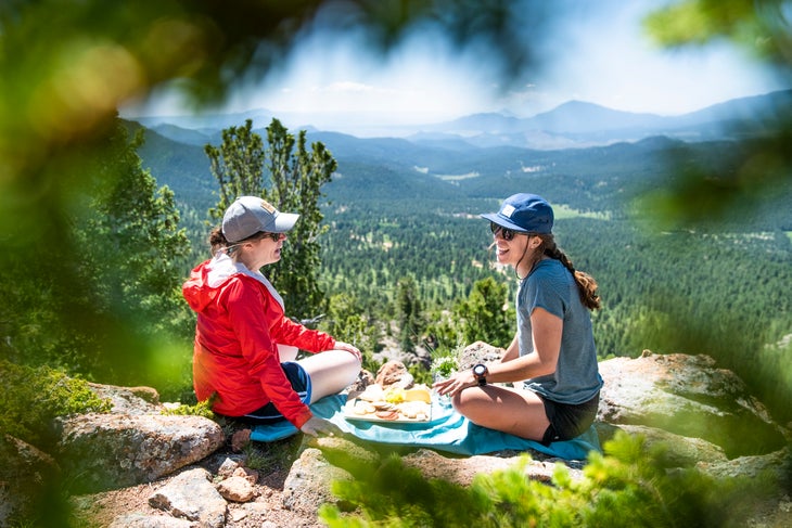 20190710_ViewRanger_273 Two white women laugh and sit on rocks eating cheese and grapes. Tree branches blur the foreground and mountains fill the background.