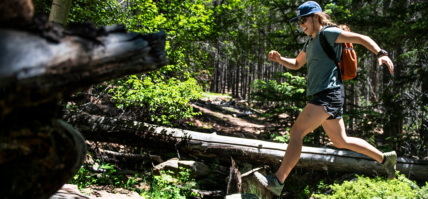 "White woman jumps across rocks in a stream while smiling."