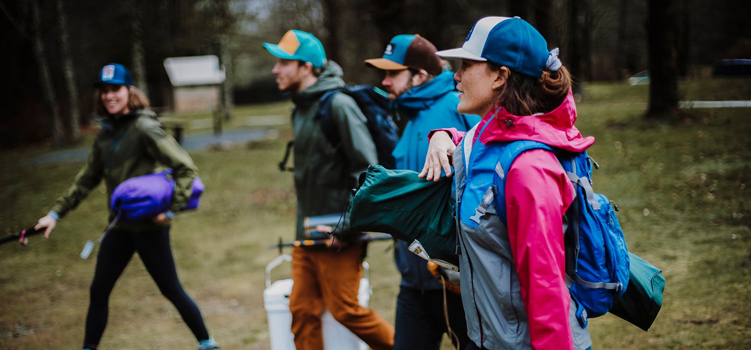 "Four young white adults hold camping gear and walk past trees in brightly colored rain Outdoor Research jackets."