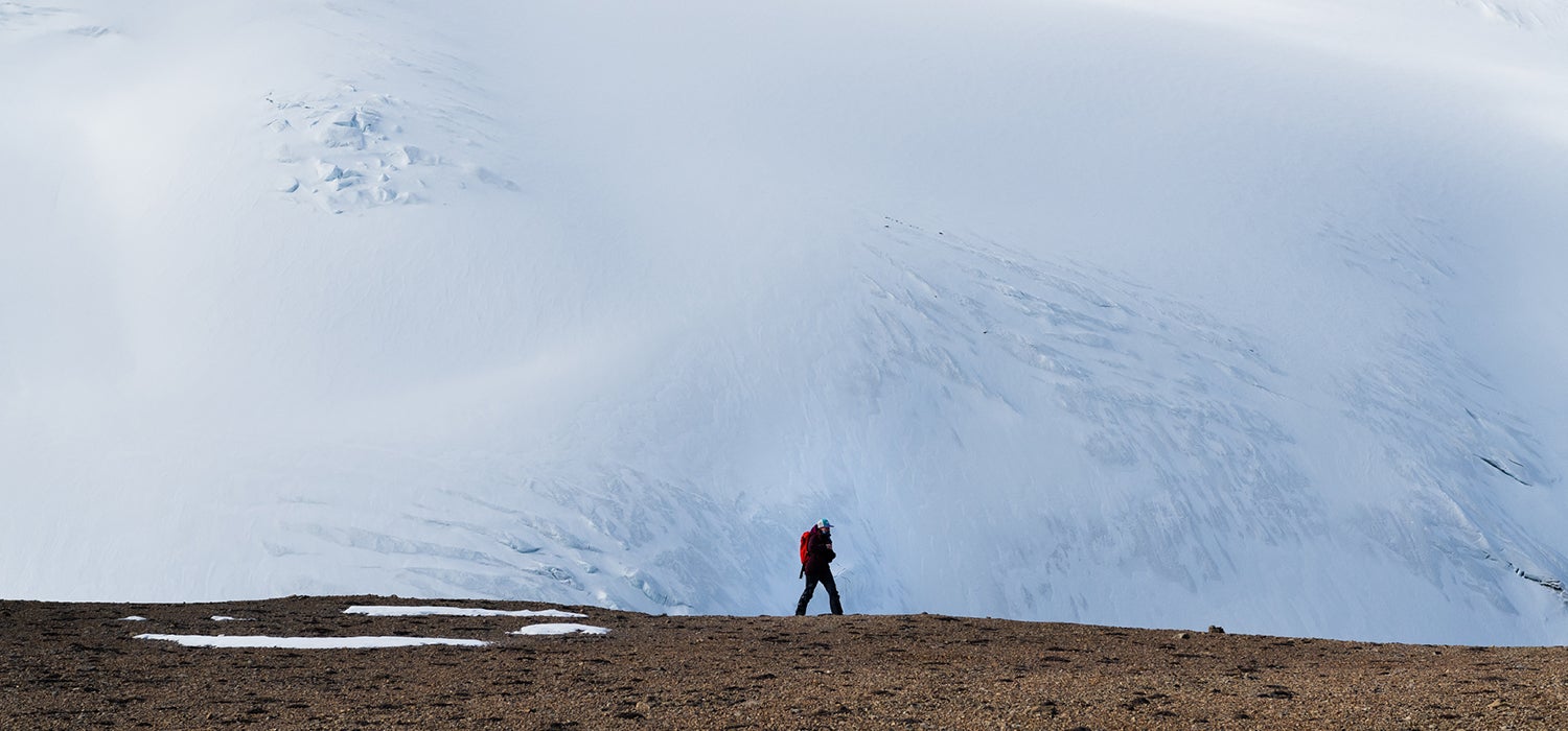 "Woman in black outfit with red backpack walks across a dirt ridge. Snowy mountainside in the background."