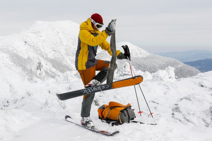 VT80OR372EMB Person in AscentShell Skyward II Jacket and pants stands on a mountain top pulling off a skin from the bottom of a ski while wearing it.