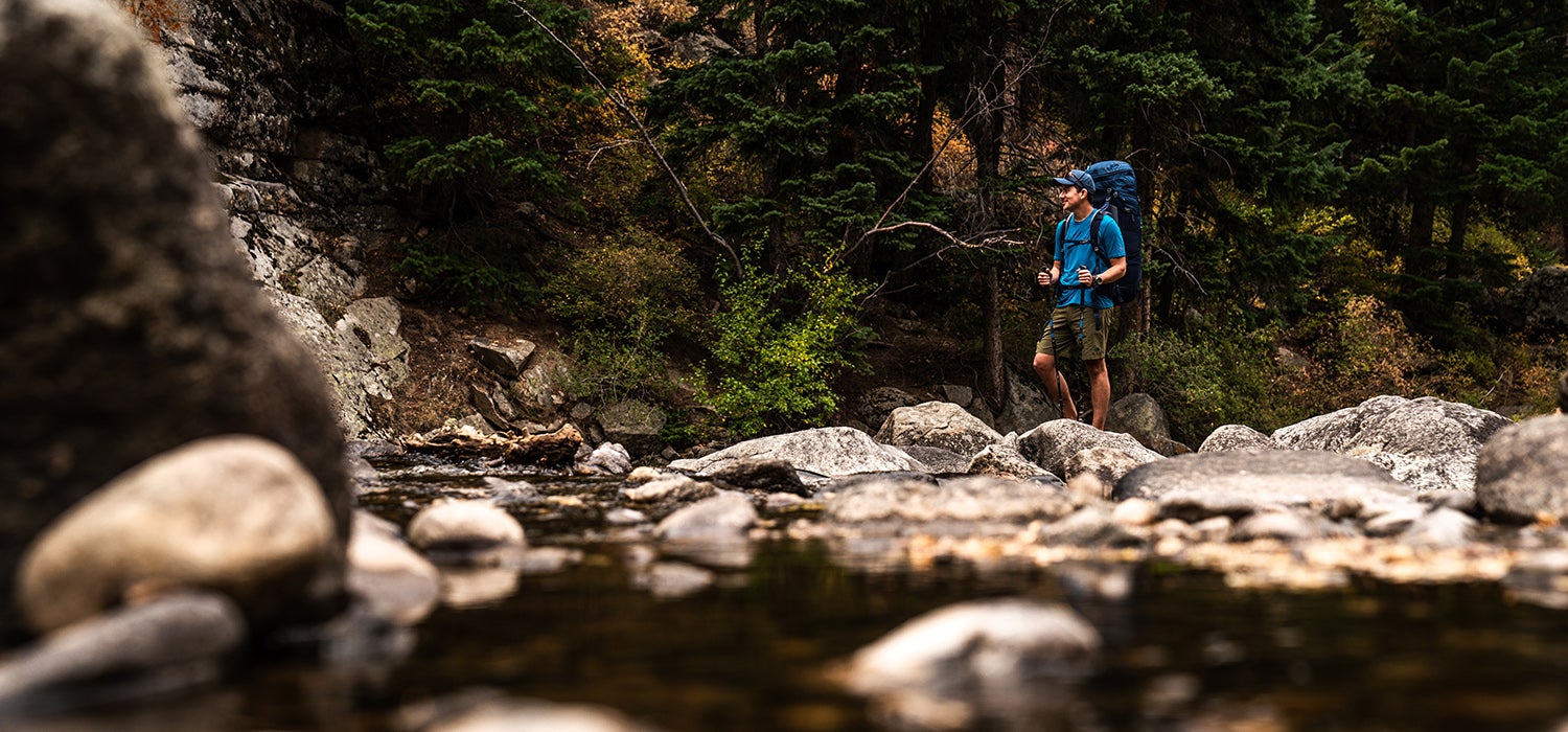 "Man in blue shirt and green shorts with a blue Deuter backpack on walks across rocks near a stream."