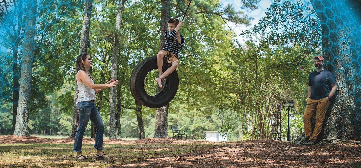 "White boy swings on tire swing toward a white woman in a white shirt and jeans. Man stands against a tree on the right side of the image."