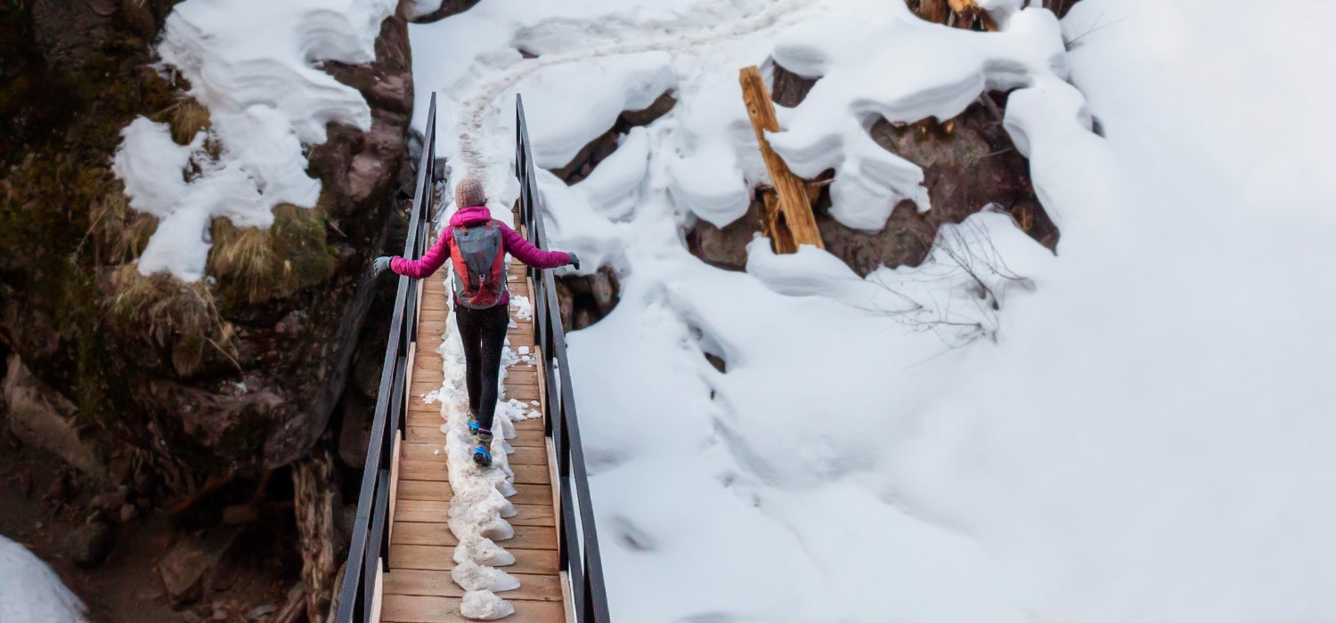 "Woman in pink jacket and black leggings walks across an icy bridge in Kahtoola MICROspikes traction."