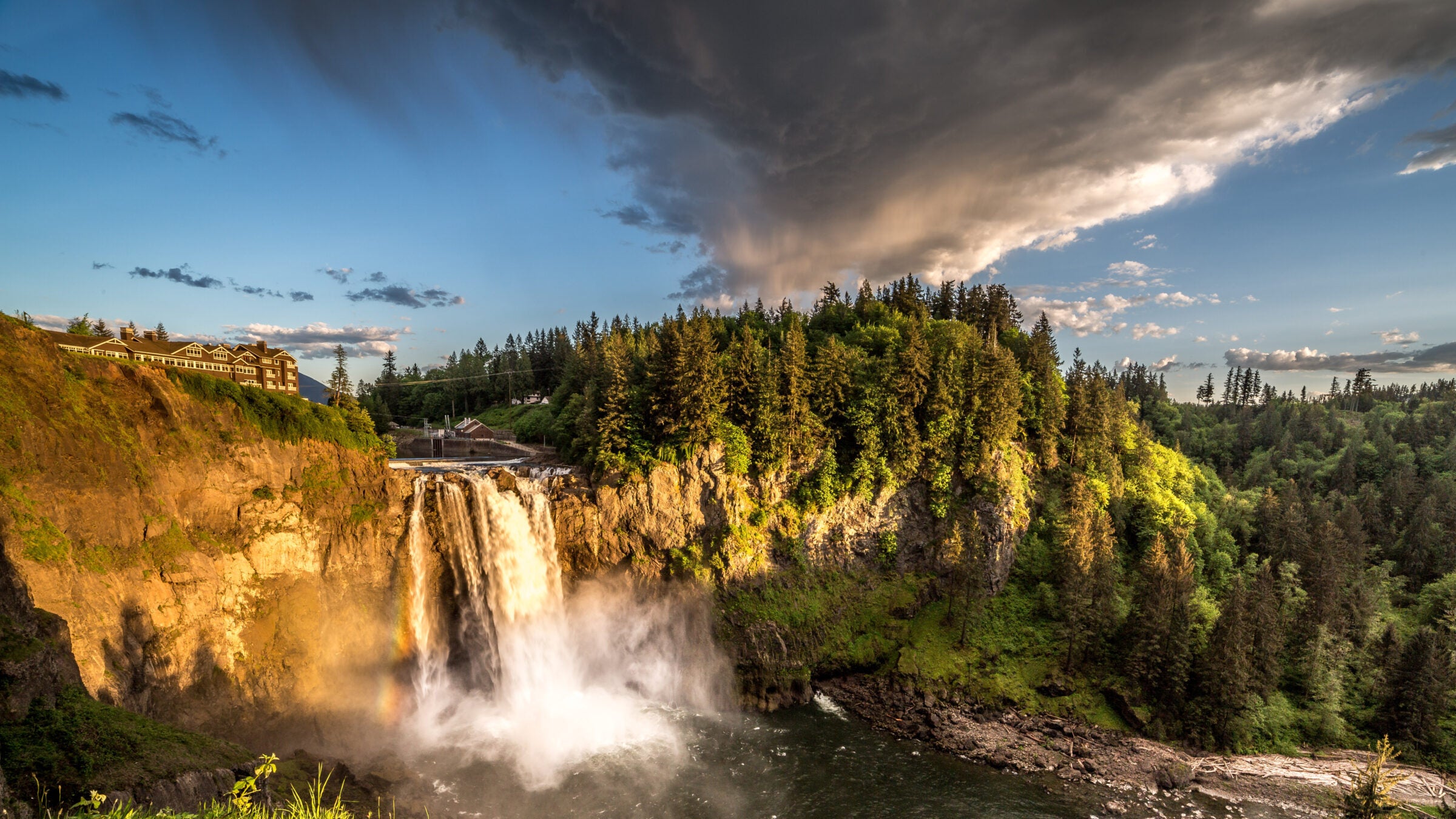 a waterfall over a large cliff between wooded hills, with a building to the left of the top of the falls