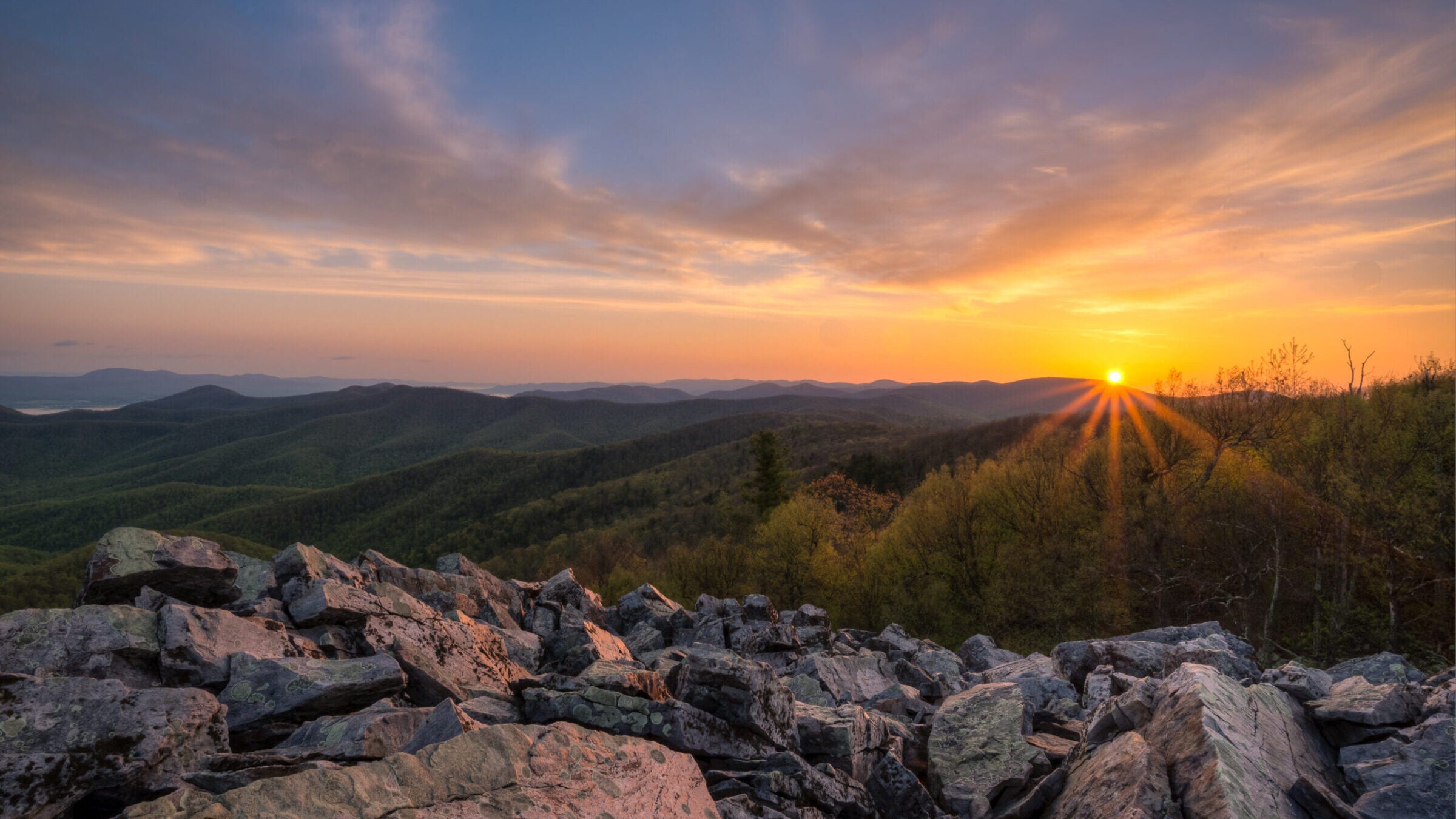 a boulder field in the foreground with rolling, forested mountains beyond; the sun is coming up over the mountains