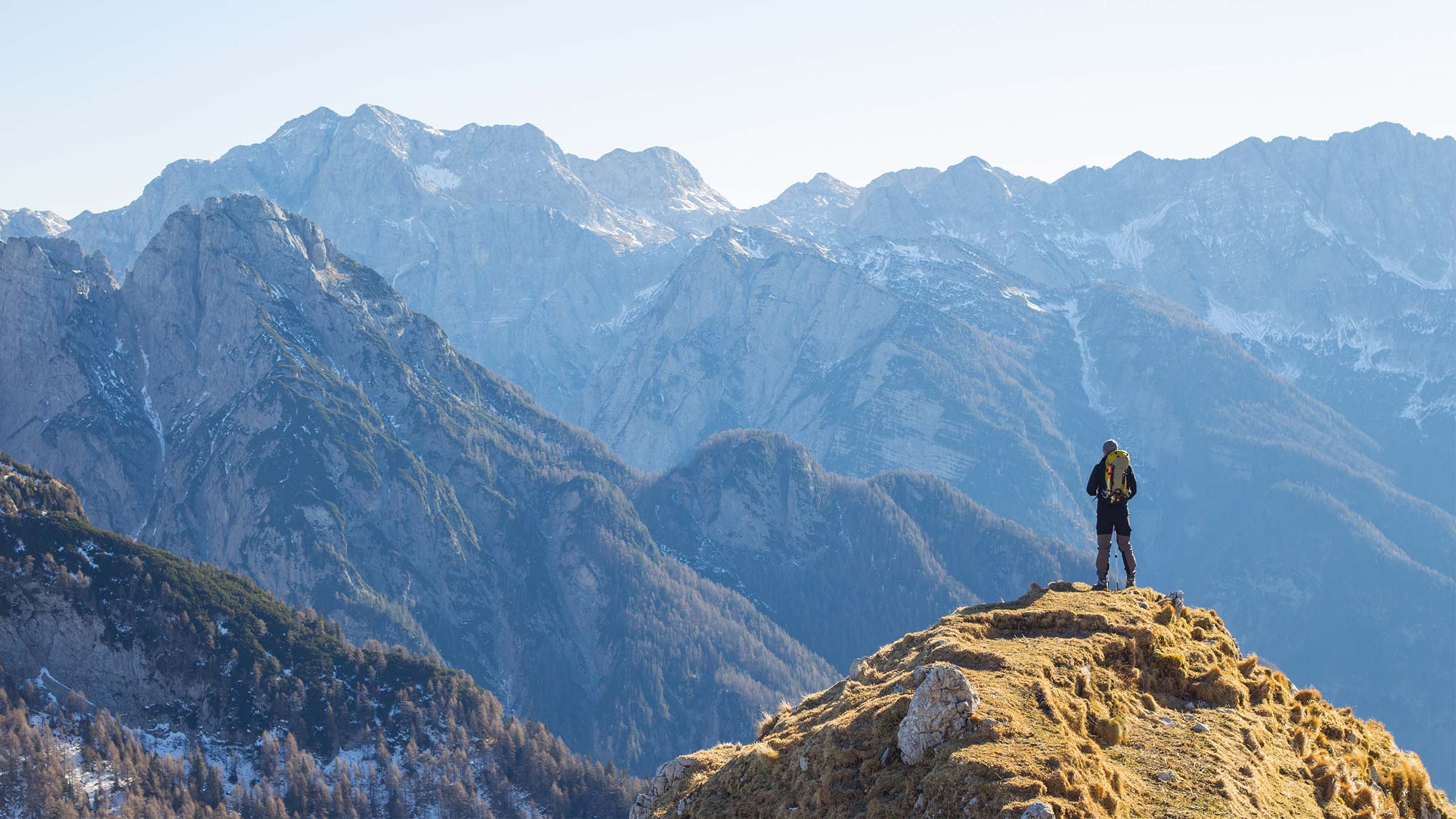 A hiker stands on a summit with high mountains in the background.
