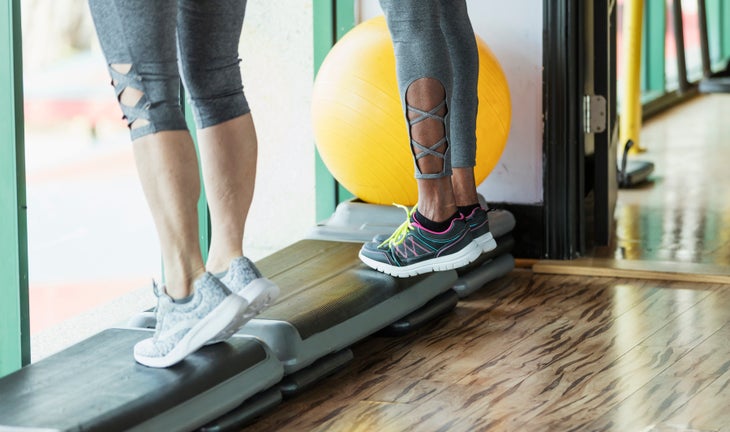 Cropped view of two women standing side by side in a gym on step exercise equipment, doing calf raises.