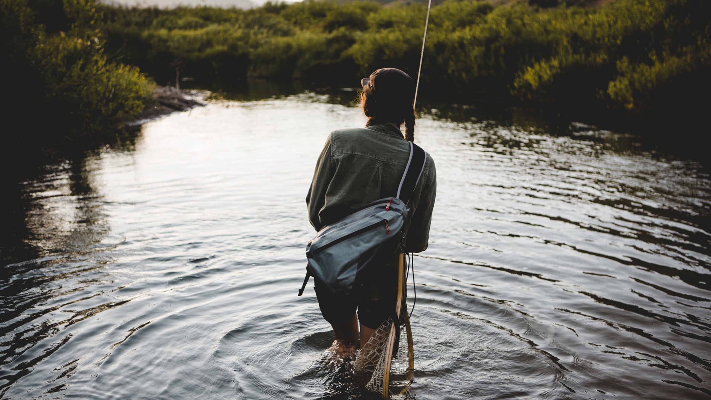 A woman fly fishing a small stream, her back is to us and she is carrying a small grey sling bag. It is sunset and there is golden light reflecting on the water.