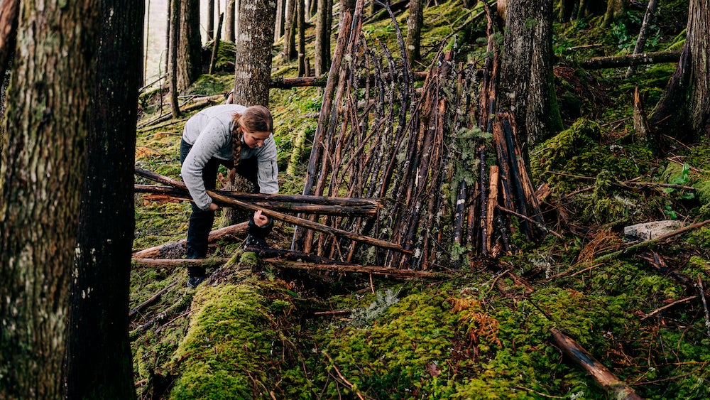 A woman gathers logs to create a survival shelter in a forested area