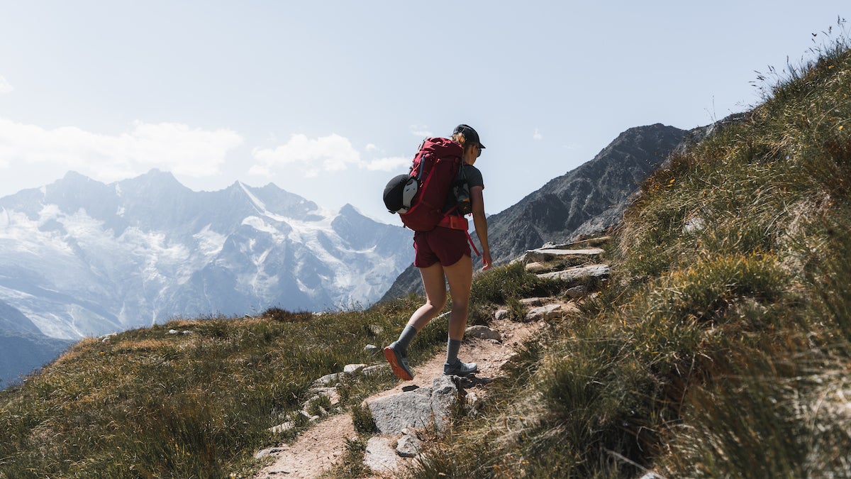 Female hiker carrying backpack walking uphill in front of mountains