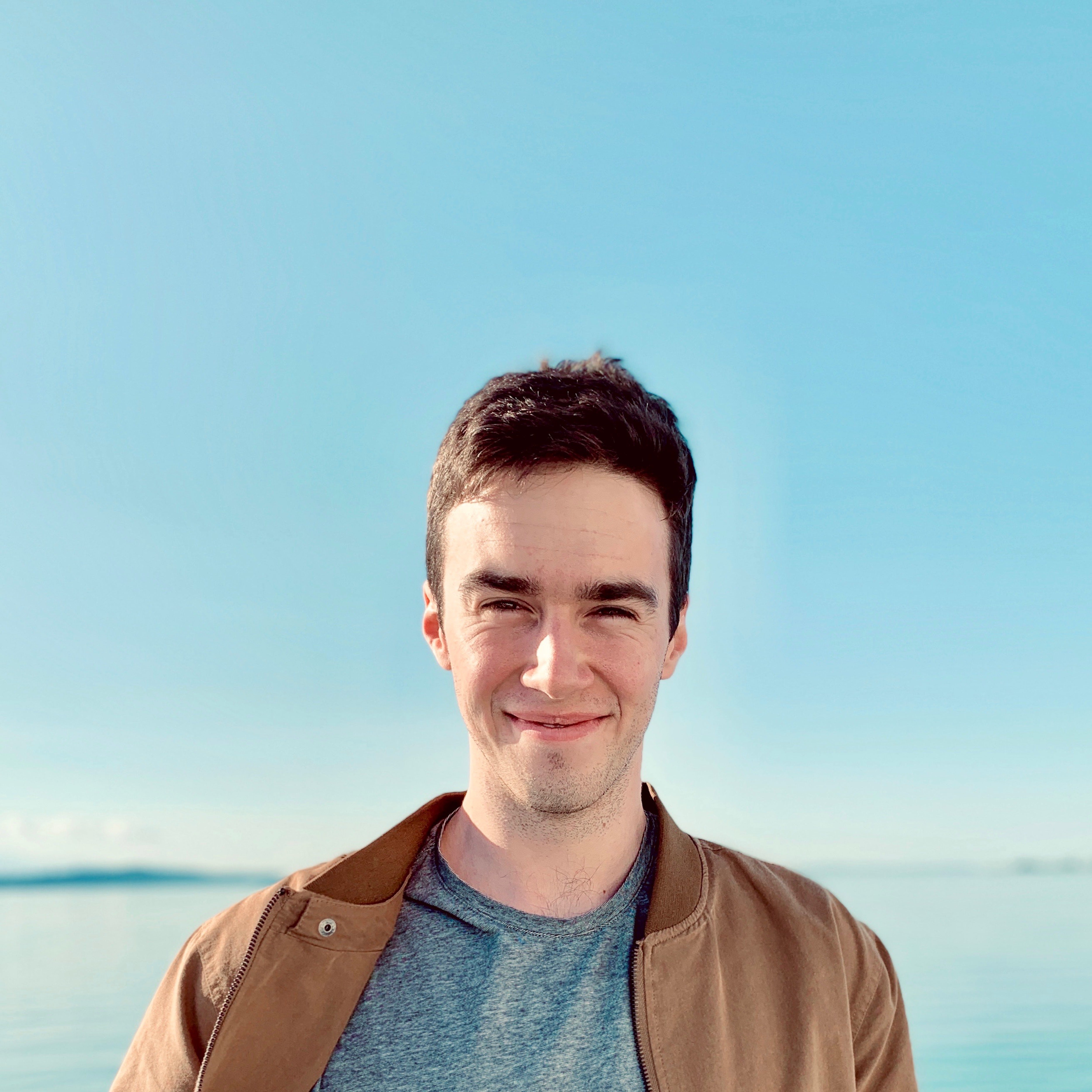 Dave bonan-headshot Dave Bonan smiles at the camera in a brown leather jacket with a clear blue sky and body of water in the background.