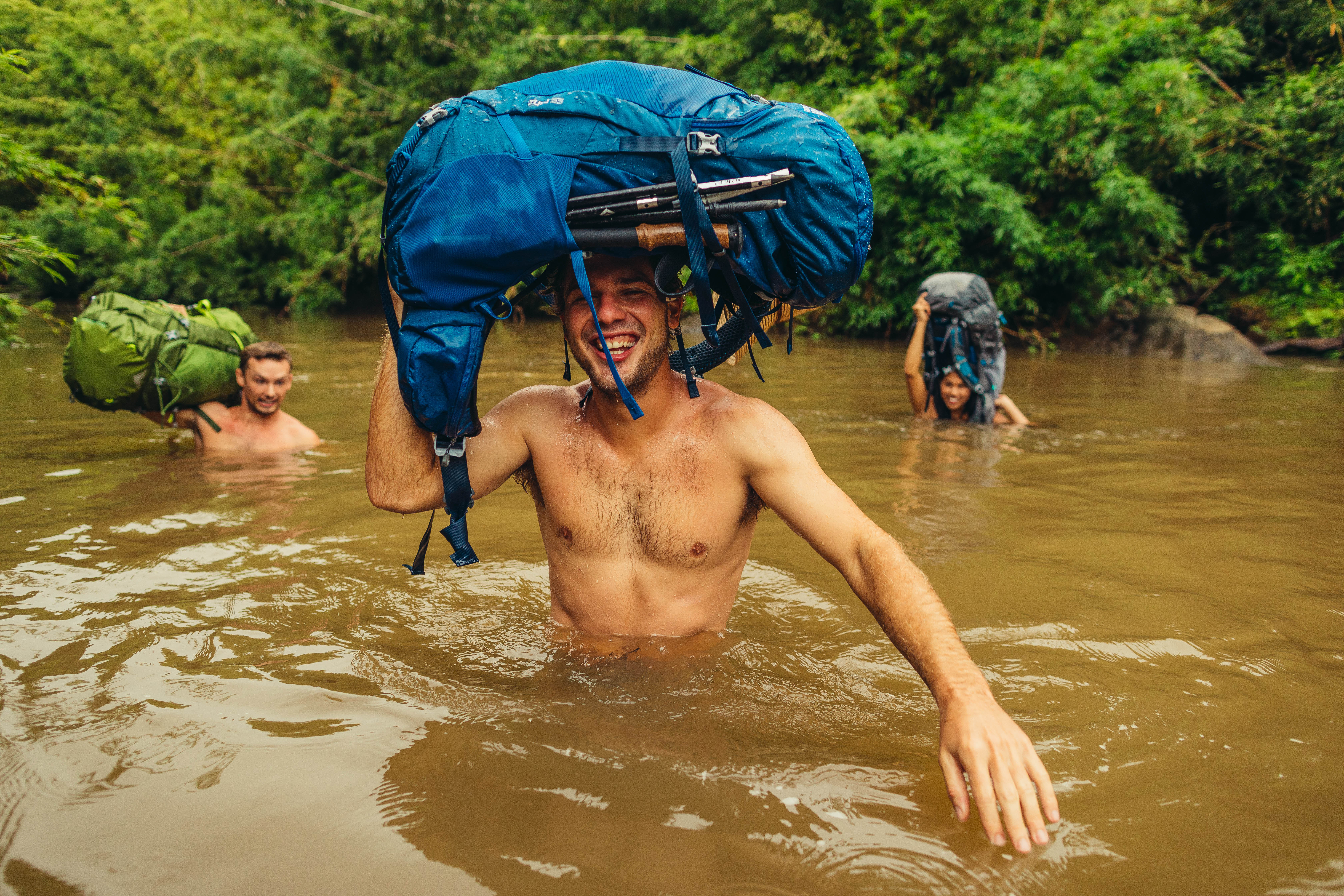 GMP_S19_ZuluJade_Hawaii-0453 Three people cross a muddy river in Hawaii laughing and holding their Gregory backpacking packs over their heads.