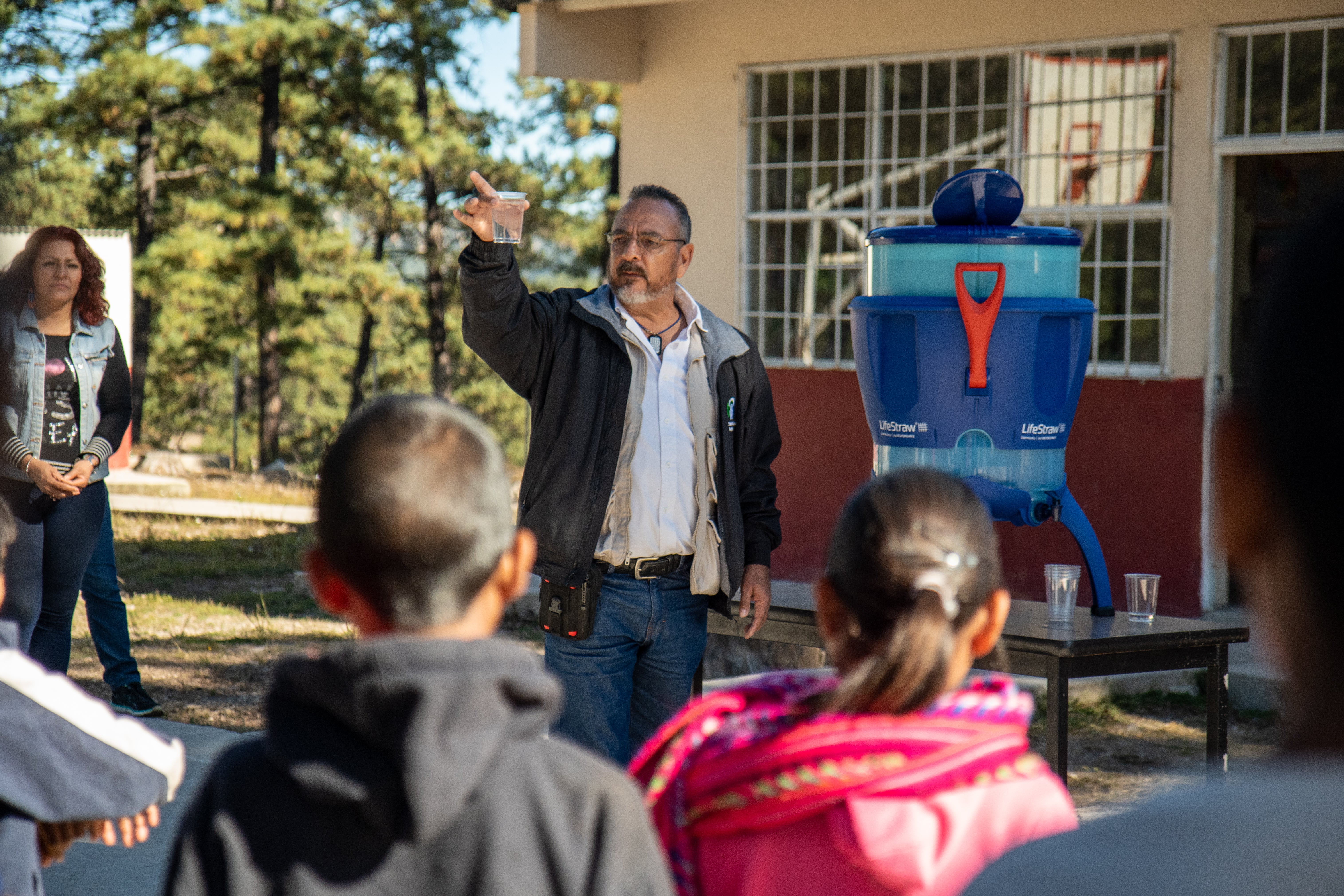 DSC09526 Department of Health official from Chihuahua holds up a glass of clear filtered water at the front of the presentation space in the courtyard by the…