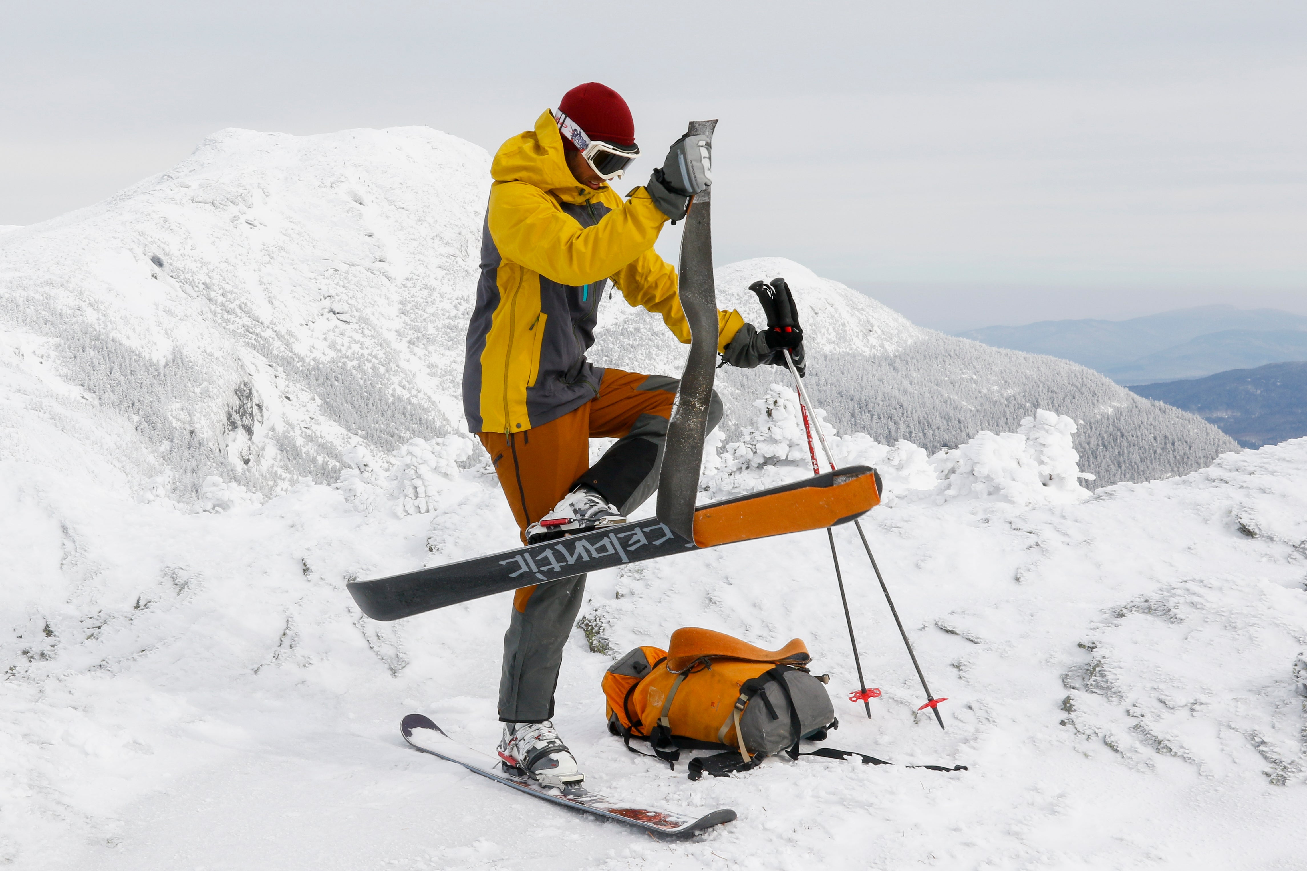 VT80OR372EMB Person in AscentShell Skyward II Jacket and pants stands on a mountain top pulling off a skin from the bottom of a ski while wearing it.